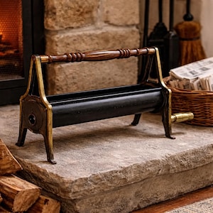 May include: A black and gold fireplace log holder with a wooden handle. The holder is on a stone hearth, with a basket of newspapers and a fireplace in the background. Firewood is stacked on the left.
