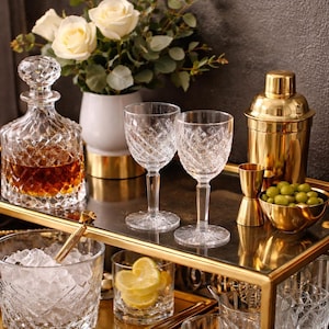 May include: A crystal decanter and two wine glasses with diamond-cut patterns sit on a gold-trimmed tray, alongside a gold cocktail shaker, jigger, and bowl of olives. A vase of white roses and greenery adds a touch of elegance to the barware setup.