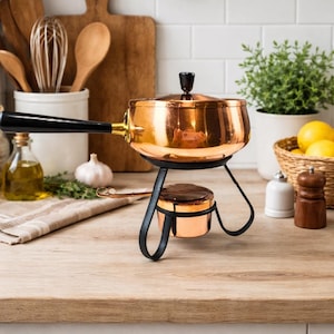 May include: A copper fondue pot with a black handle and stand. The pot has a lid with a black knob. The stand is black and has a small burner underneath. The pot is sitting on a wooden surface.
