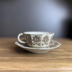 May include: Cream-colored teacup and saucer set. The cup and saucer have a detailed embossed floral design in a darker cream color. The cup has a handle and sits on a matching saucer, both placed on a wooden surface.