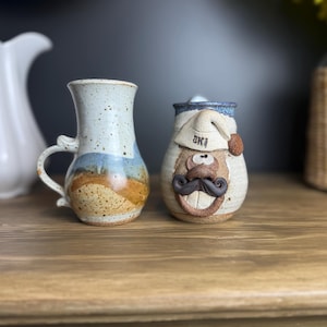 Puede incluir: Dos tazas de cerámica sobre una superficie de madera. Una es una jarra beige y azul moteada con un asa. La otra es una taza caprichosa con una cara, un bigote y un sombrero que dice "SKI".