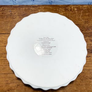 May include: A white round ceramic dish with a scalloped edge. The underside features a printed label with the text Villeroy & Boch, Vilbofour, Botanica, and instructions including dishwasher-safe, microwave-safe, and freezer-to-oven.