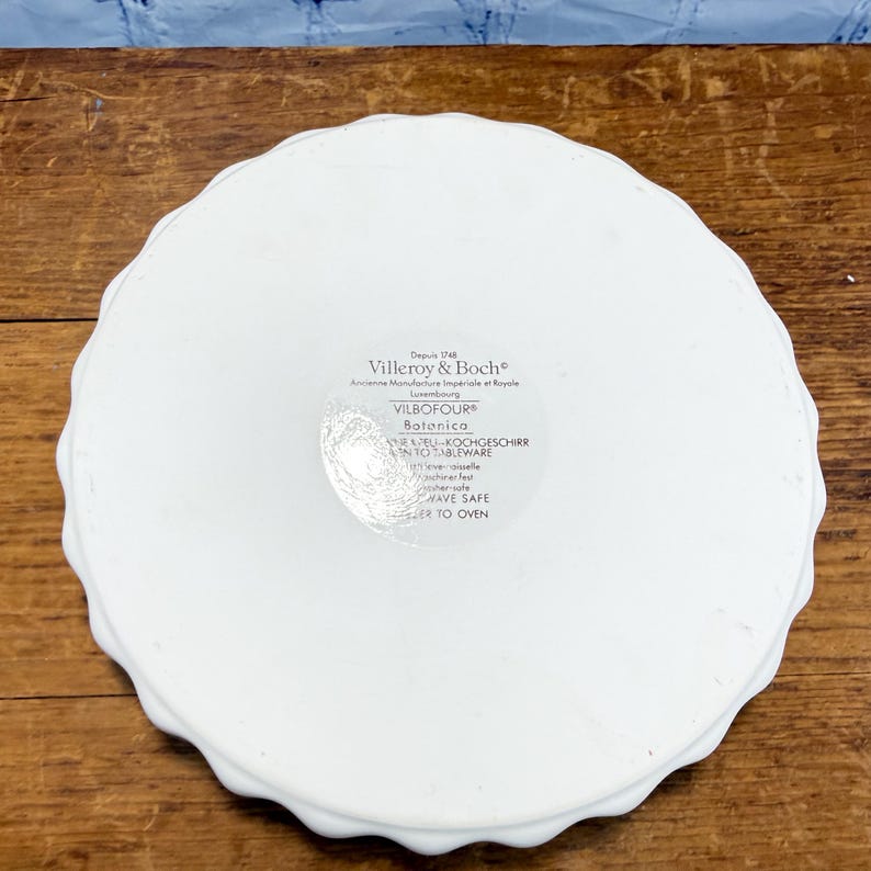 May include: A white round ceramic dish with a scalloped edge. The underside features a printed label with the text Villeroy & Boch, Vilbofour, Botanica, and instructions including dishwasher-safe, microwave-safe, and freezer-to-oven.