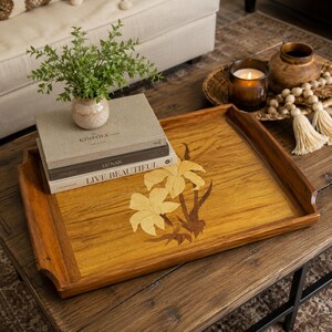 May include: A rectangular wooden serving tray featuring a light-colored wood inlay of two flowers in the center. The tray has raised edges and a natural brown wood grain finish. The inlay displays two blossoms with stems in contrasting wood tones.