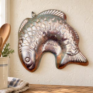 Könnte beinhalten: Eine metallische fischförmige Backform mit kupfer- und silberfarbenem Finish, die detaillierte geprägte Schuppen, Flossen und ein kreisförmiges Auge aufweist. Die Form enthält eine kleine Metallöse an der Oberseite zum Aufhängen.