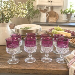 May include: A set of six stemmed goblets featuring deep magenta glass bowls with a textured, clear glass base. Each glass has a rounded, faceted pattern on the lower portion and a smooth, vibrant magenta upper rim, arranged on a wooden surface.
