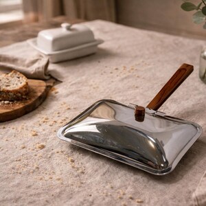 May include: A shiny, rectangular crumb sweeper with a wooden handle rests on a beige tablecloth. A white butter dish and a loaf of bread are also on the table. A small glass vase with greenery is in the background.