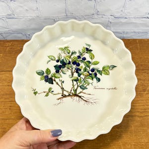 May include: A round, cream-colored ceramic fluted tart dish featuring a botanical illustration of a Vaccinium myrtillus blueberry plant in the center. The design displays green leaves, dark blue berries, and visible roots on a glossy surface.