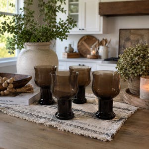 May include: A set of four smoke-colored glass goblets with a rounded bowl and a thick, sturdy base. The glasses are arranged on a rectangular, cream-colored woven fabric runner with black striped details and fringed edges on the short sides.