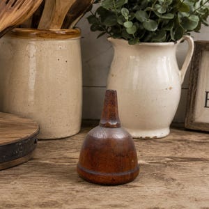 May include: A small, dark brown wooden funnel with a smooth, rounded body and a tapered spout. The funnel features a visible wood grain texture and a simple, functional design, standing upright as a standalone object in the center of the frame.