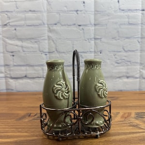 May include: A salt and pepper shaker set. The set includes two olive green ceramic shakers with a floral design, held in a decorative metal holder with a handle. The shakers sit on a wooden surface against a white brick wall.