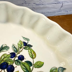 May include: A white ceramic pie dish with a scalloped edge and a painted botanical illustration of dark blue blueberries and green leaves on the interior surface. The dish features a smooth, glossy finish and a curved, fluted rim design.