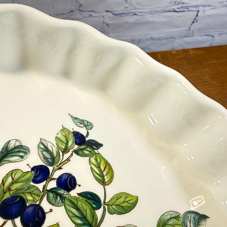May include: A white ceramic pie dish with a scalloped edge and a painted botanical illustration of dark blue blueberries and green leaves on the interior surface. The dish features a smooth, glossy finish and a curved, fluted rim design.