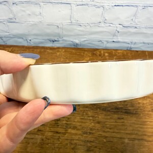 May include: A round, off-white ceramic dish with a scalloped edge and a thin, dark brown rim. The dish features a smooth, glossy finish on the exterior side walls and a shallow, circular form, showing a simple and functional design for serving.