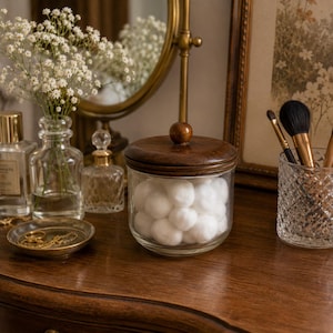 May include: A glass jar with a wooden lid filled with cotton balls sits on a wooden surface. A small gold-colored tray with jewelry, a vase of flowers, and a decorative mirror are also visible.