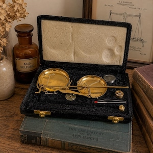 May include: A vintage brass balance scale set housed in a black velvet-lined case. The set includes two circular brass pans suspended by thin chains, a central beam, small metal weights, and a pair of tweezers, all arranged in molded compartments.