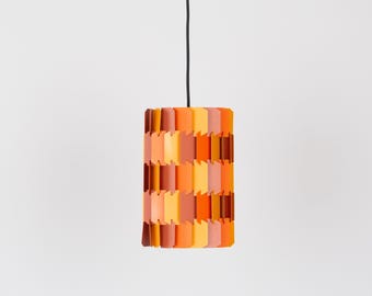 May include: A cylindrical pendant light fixture with a colorful, layered design. The light features a series of rectangular panels in shades of orange, yellow, and brown. The light hangs from a black cord against a white background.