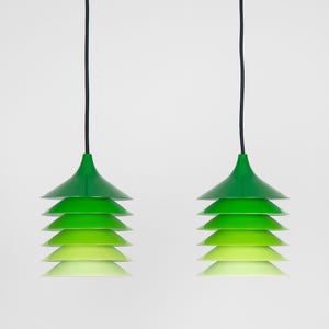 May include: Two modern pendant lights with a layered, tiered design. Each light features a series of green shades that graduate in color from dark to light. The lights hang from black cords against a white background.