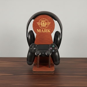 May include: A wooden headphone and controller stand with the text "BEST DAD EVER" and the name "MARK" in gold lettering. A black gaming controller and headphones are displayed on the stand, which is set on a wooden surface.
