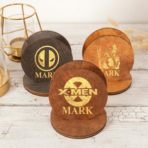May include: Wooden coasters with gold lettering and superhero designs. One coaster features a Deadpool logo, another an X-Men logo, and a third a superhero figure. Each coaster is round and sits in a matching wooden holder. The word "MARK" is below the logos.
