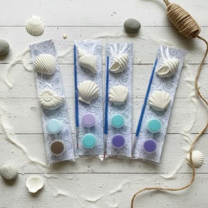 May include: Three craft kits in clear plastic packaging, each containing two white plaster seashell figures, a blue paintbrush, and two small circular paint pots in shades of purple, blue, or brown, set on a seashell-patterned card backing.