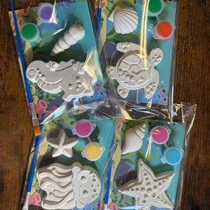 May include: Four clear plastic bags containing craft kits with white plaster sea creature figures, including a seahorse, turtle, starfish, and jellyfish. Each kit includes a blue paintbrush and small containers of colorful acrylic paint.