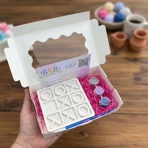 May include: A white cardboard box containing a tic-tac-toe game board, three small paint pots in gray, purple, and blue, and a blue paintbrush. The box includes a B&B Handcrafted card with a QR code and pink shredded paper filler.