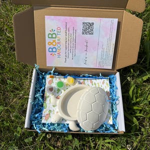 May include: A craft kit in a cardboard box containing a white ceramic turtle figurine with a removable shell, a small paintbrush, two small paint pots, and a printed instruction card with the text B&B HANDCRAFTED and a QR code, all nestled in blue paper filler.