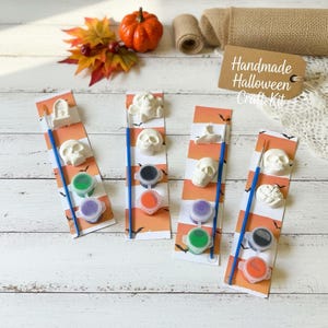May include: Four vertical cardboard strips with orange and white stripes and black bat silhouettes. Each strip includes a small white plaster figure, two small paint pots, and a blue paintbrush. A tag reads Handmade Halloween Craft Kit.