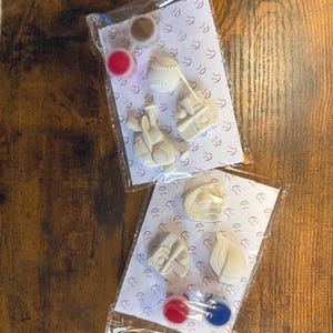 May include: Two clear plastic bags containing white baseball-themed plaster figures, small paint pots in red, blue, and tan, and a thin paintbrush. Each bag includes a printed card with a repeating baseball pattern as a backing for the items.
