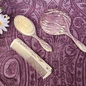 May include: A vintage vanity set featuring a silver-toned hand mirror, hairbrush, and comb. The set is displayed on a purple paisley patterned fabric. The mirror and comb have decorative silver accents.