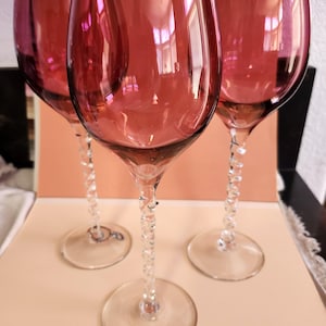 May include: Three wine glasses with deep cranberry-colored bowls and clear, twisted glass stems. Each glass features a smooth, rounded rim and a circular base, showcasing a consistent design across the set with transparent, spiraled stems.