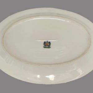 Pu&ograve; includere: Un piatto da portata ovale in ceramica bianca. Il lato inferiore presenta un emblema decorativo con accenti blu, oro e verde. Il piatto ha un bordo leggermente rialzato e una superficie liscia e lucida.