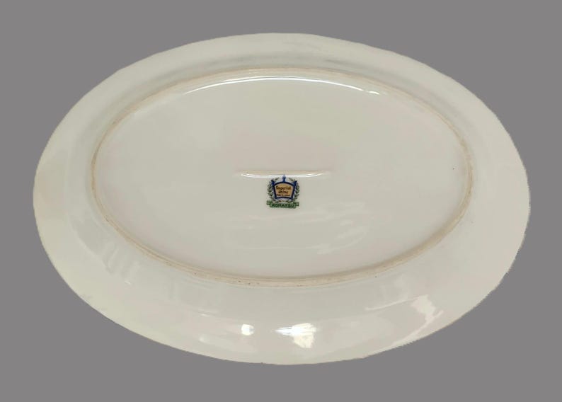 Pu&ograve; includere: Un piatto da portata ovale in ceramica bianca. Il lato inferiore presenta un emblema decorativo con accenti blu, oro e verde. Il piatto ha un bordo leggermente rialzato e una superficie liscia e lucida.