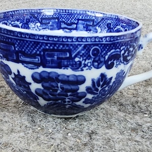 Peut inclure: Tasse à thé en céramique bleue et blanche avec une anse. La tasse présente un motif Willow détaillé, avec des scènes de pagodes, de ponts et de feuillages. La tasse est posée sur un fond neutre.