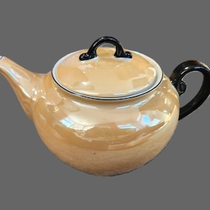 May include: A pearlescent, peach-coloured teapot with a black handle and lid handle. The teapot has a rounded body, a curved spout, and a lid. The rim of the lid and the base of the spout are trimmed with a thin white line. A classic tea accessory.