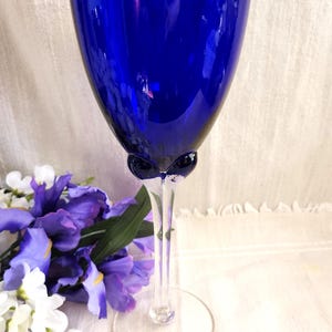 May include: A tall, elegant cobalt blue wine glass with a clear stem and base. The glass has a rounded bowl and a delicate, elongated stem. The glass is next to a bouquet of purple and white flowers, creating a visually appealing contrast.