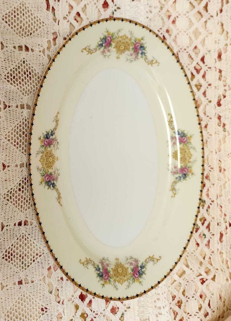 Pu&ograve; includere: Piatto da portata ovale color crema con un motivo floreale. Il piatto presenta un bordo dorato con accenti neri e un'area ovale centrale. Composizioni floreali in rosa, blu e giallo adornano i bordi.