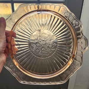 May include: A vintage, pink glass serving tray with a floral design in the center and along the scalloped edge. The tray has a radial pattern emanating from the center and two small handles. The tray is held by a hand.