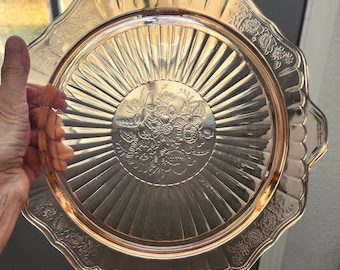Puede incluir: Una bandeja de servir de cristal rosa vintage con un diseño floral en el centro y a lo largo del borde festoneado. La bandeja tiene un patrón radial que emana del centro y dos asas pequeñas. La bandeja está sujeta por una mano.