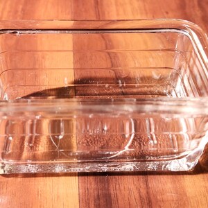 May include: A clear, rectangular glass dish with a slightly textured surface. The dish has a square shape with a slightly raised rim and is sitting on a wooden surface. The dish is designed for food storage or serving.