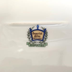 Pu&ograve; includere: Un piatto di porcellana bianca con un emblema decorativo. L'emblema presenta le parole "Imperial China Made in Japan" all'interno di uno stemma blu e oro, circondato da foglie di alloro. La parola "KOMATSU" &egrave; sotto.