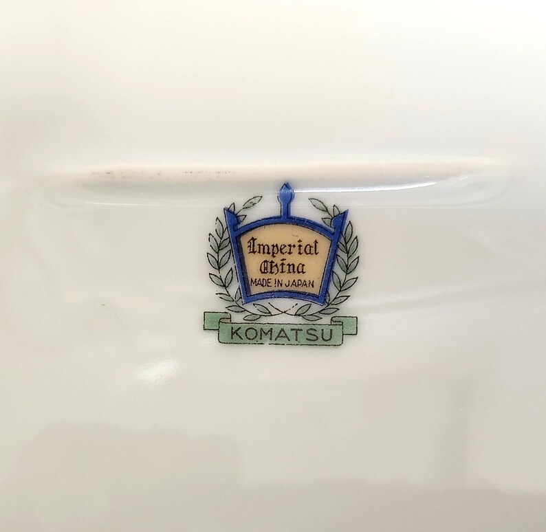 Pu&ograve; includere: Un piatto di porcellana bianca con un emblema decorativo. L'emblema presenta le parole "Imperial China Made in Japan" all'interno di uno stemma blu e oro, circondato da foglie di alloro. La parola "KOMATSU" &egrave; sotto.