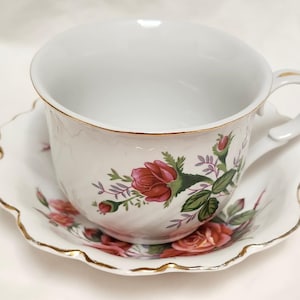 May include: White teacup and saucer set with a floral rose design. The teacup and saucer have a scalloped edge with gold trim. The roses are red and pink with green leaves and purple accents. The teacup has a curved handle.
