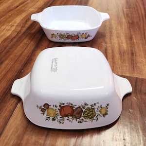 May include: Two white square Corning Ware baking dishes with handles. The dishes have a colorful fruit and vegetable pattern around the bottom edge. One dish is upright, and the other is upside down, showing the bottom.