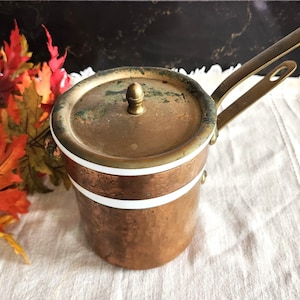 May include: A cylindrical copper pot with a matching lid, featuring a brass handle and a white enamel band around the upper section. The metal surfaces show signs of oxidation and wear, with a small brass knob centered on the lid.