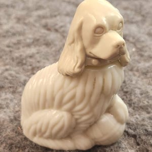 Puede incluir: Figurilla de color crema de un perro sentado, posiblemente un Cocker Spaniel, con textura de pelaje detallada. El perro tiene orejas caídas, un pequeño collar y una expresión suave. La figurilla está hecha de un material sólido, probablemente cerámica o resina.