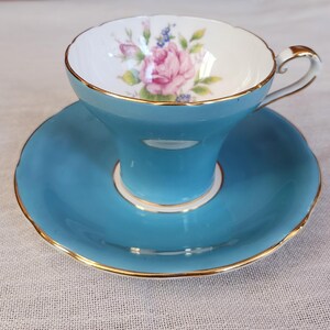 May include: A vintage teacup and saucer set. The teacup is a vibrant turquoise with a gold rim and handle. The interior of the cup features a pink rose floral design. The matching saucer has a scalloped edge and gold trim, creating an elegant aesthetic.