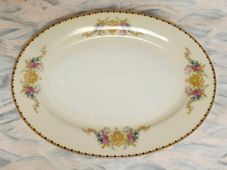 Pu&ograve; includere: Piatto ovale da portata color crema con bordo dorato e nero. Il piatto presenta disegni floreali in rosa, blu e giallo. I disegni floreali sono posizionati alle estremit&agrave; e al centro del piatto, aggiungendo un tocco decorativo.