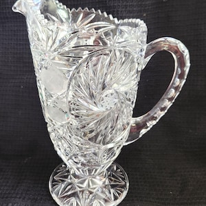 May include: A clear, cut-glass jug with a handle and spout. The jug features intricate starburst and floral patterns, with a pedestal base. The handle is adorned with small, rounded details. The jug is approximately 20 cm tall.